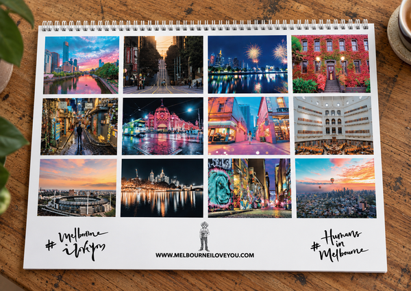A spiral-bound wall calendar or print laid flat on a wooden table, featuring a grid of colorful Melbourne cityscape photographs with text reading '#Welcome to Melbourne' and 'Humans love Melbourne' plus the website MELBOURNEILOVEYOU.COM.