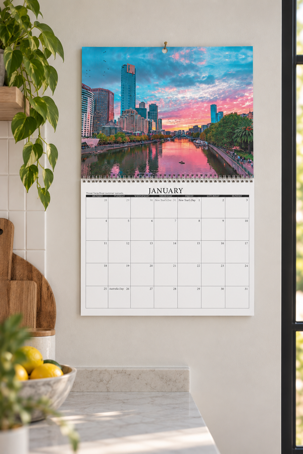 A January wall calendar hanging on a light beige wall in a kitchen setting, featuring a colorful city skyline sunset print on the top half and a white monthly grid on the bottom half.